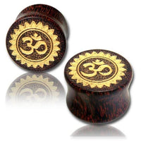 Load image into Gallery viewer, Two round Holz Plug Tamarind Wood Tribal Tunnels feature hand-carved and gold-painted Om designs with a laser-engraved sunburst, displayed on a white background with subtle reflection.
