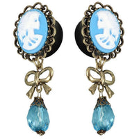 Cargar imagen en el visor de la galería, Vintage-style earrings: blue and white acrylic cameo skeleton woman, gold brass border, bow accent, faceted blue glass teardrop pendants—Plug Piercing.
