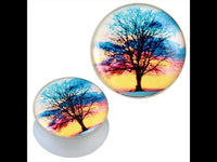 Load image into Gallery viewer, Two round white acrylic plugs feature a black tree silhouette against a colorful sunset in blue, pink, orange, and yellow. These glossy Piercing Plugs are called &quot;Plug weiß Acryl Baum schwarz Sonnenuntergang bunt Ohrschmuck.
