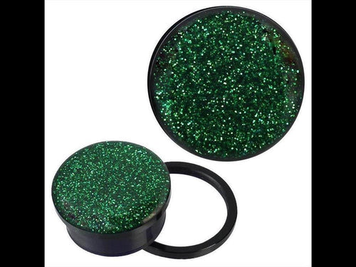 A pair of Schraub Plug Tunnel Acryl schwarz grüner Glitzer Piercing Ohrschmuck rests on a white background. One plug stands upright, displaying its green glitter top, while the other lies on its side with a black ring next to it.