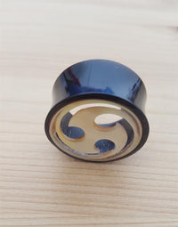 Load image into Gallery viewer, A glossy black Horn Plug Tunnel Perlmutt Tomoe Inlay weiß double flared with a beige, three-swirl design rests on a light wooden surface, serving as decorative tribal piercing jewelry.
