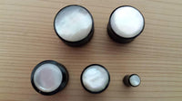 Cargar imagen en el visor de la galería, Five Buffalo Horn Plug Perlmutt Inlay weiß handgeschnitzt Lobe Piercing Tunnels, arranged on a light wooden surface, feature shiny white tops and differ in size, with one tunnel significantly smaller than the rest.
