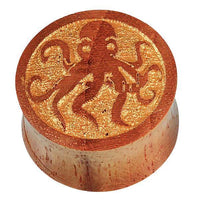 Load image into Gallery viewer, The Teakholz Plug braun gold bemalt Oktopus double flared features a round wooden design with a polished finish and a detailed octopus engraving on top, highlighting its intricate craftsmanship.
