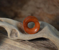 Load image into Gallery viewer, A Blutholz Flesh Tunnel rötlich, handmade and double flared, with a smooth round shape and central hole, rests on pale driftwood against a blurred brown background.
