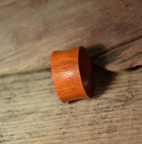 Load image into Gallery viewer, A smooth, wide Blutholz Flesh Tunnel rötlich, handmade and double flared, with a natural brown finish rests on rustic wood—an elegant choice for organic piercing enthusiasts.
