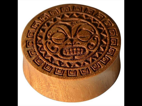 The Holz Teakholz Plug Sonnen Maske double flared is a round wooden plug with an intricate, stylized grinning face and geometric patterns inspired by ancient Mesoamerican or Aztec motifs.