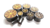 Load image into Gallery viewer, A set of Plug Arangholz Wikinger Kompass Inlay double flared plugs in black, each with a golden Icelandic Vegvisir symbol on the front, displayed on a white background—ideal for fans of unique Holzplug style and Arangholz craftsmanship.
