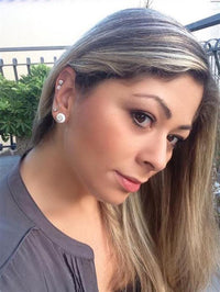 Load image into Gallery viewer, A woman with long, straight blonde hair wearing a grey top looks at the camera outdoors, greenery and a metal fence behind her. She is wearing multiple Ohstecker Shiva Auge rund 925 Silber earrings in sizes 6mm, 10mm, and 12mm.
