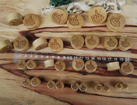 Load image into Gallery viewer, A set of round wooden Krokodilholz Plug stamps, hellbraun gemasert, with Skeletthand Mittelfinger Lasergravur-Tribal designs, arranged above and below a clear plastic ruler on a wooden surface. The stamps vary in size.
