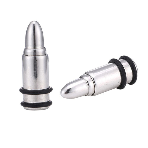 Two silberne Edelstahl Plugs in Patronenhülsenform aus Chirurgenstahl mit schwarzen Gummiringen—einer stehend, einer liegend—werden auf weißem Hintergrund gezeigt.