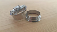 Load image into Gallery viewer, Two silver rings on a light wooden surface: one features an ornate faceted stone with wing accents like the Edelstahl Tunnel Plug Flügel Herz Stein, while the other is plain and smooth.
