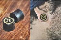 Load image into Gallery viewer, A close-up of an ear wearing a black gauge with a gold Om symbol. Next to it, two matching Holz Plug Tamarind Wood Blume Om gold, hand-carved and laser-engraved tribal tunnels, are displayed on wood.

