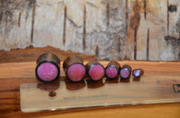 Cargar imagen en el visor de la galería, Six Holz Plug Sonoholz rosa schillernd Resin Inlay double flared plugs in graduated sizes, with pink centers, are lined up beside a transparent ruler on a wood-textured surface.
