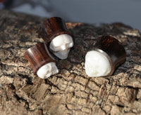 Load image into Gallery viewer, Three small Holz Plug Sonoholz Bone Inlay Totenschädel double flared plugs with carved skull faces rest on tree bark, sunlight revealing their details. These unique pieces bring organic style to any Ohrschmuck collection.
