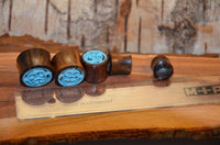 Cargar imagen en el visor de la galería, The Tunnel Sonoholz Resin Türkis Inlay Spiralbögen, along with two other wooden plugs, are arranged beside a clear ruler on a rustic wooden surface.
