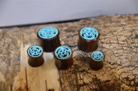 Cargar imagen en el visor de la galería, Five wooden ear plugs, including the Tunnel Sonoholz Resin Türkis Inlay Spiralbögen with elegant blue spiral designs, are shown on textured bark. The plugs vary in size and are artfully grouped for display.
