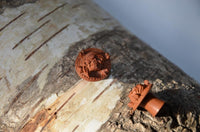 Load image into Gallery viewer, A close-up of the Holz Plug Pirat Totenkopf handgeschnitzt rotbraun double flared on rough tree bark, with another broken plug nearby in a softly lit background.
