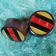 Cargar imagen en el visor de la galería, Two circular Holz Plug Tamarindenholz Resin Perlmutt Koralle Streifen Tunnel Tribal Expander plugs with dark brown sides and striped tops in red, yellow, and black rest on a green textured surface.
